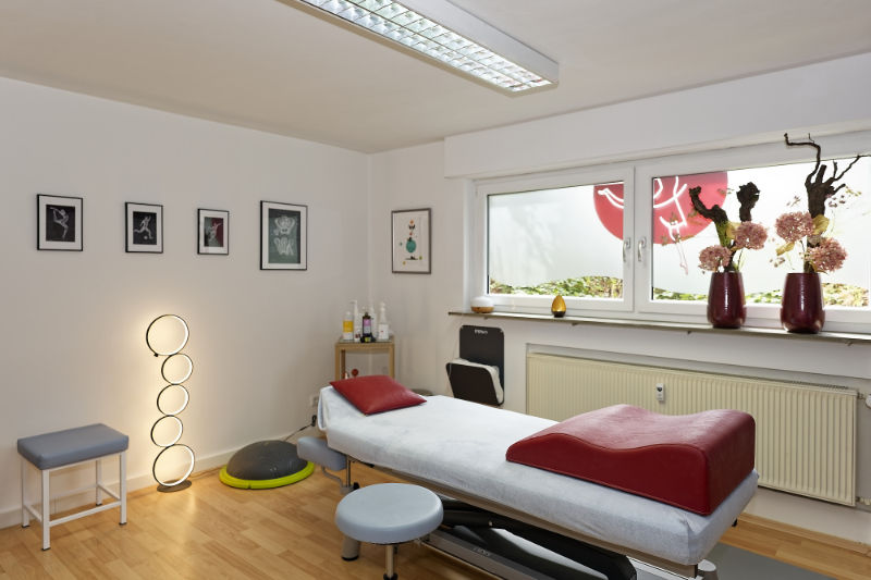 Moderner Behandlungsraum bei der Physiotherapie Oberasbach mit verstellbarem Behandlungstisch, einzigartiger runder Stehlampe, gerahmter Kunst an der Wand und dekorativen Vasen auf der Fensterbank.