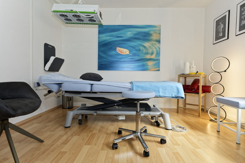 Ein Behandlungsraum mit einem verstellbaren Bett, Stuhl, medizinischer Ausrüstung und einem großen Wandbild mit einem Blatt auf dem Wasser.