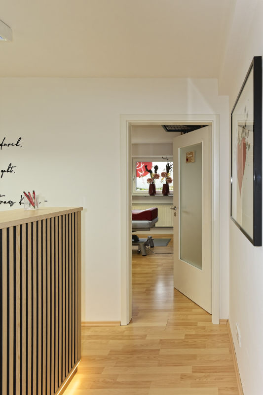 Ein Flur mit hellem Holzboden führt zu einer offenen Tür, hinter der sich ein Behandlungsraum mit Liege und großen dekorativen roten Blumen am Fenster. Am Empfang links im Bildausschnitt vereinbaren wir Ihren nächsten Physiotherapie-Termin.