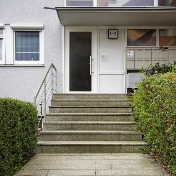 Eingang zu einem Mehrfamilienhaus mit Glastür, Stufen, Briefkästen auf der rechten Seite und Büschen am Gehweg – perfekt für Ihren nächsten Physiotherapie-Termin.