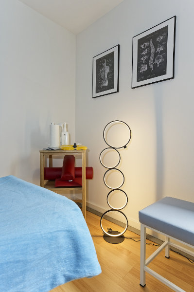 Ein minimalistischer Therapieraum für Physiotherapie-Behandlungen mit einer blau bezogenen Liege, einer modernen Ringlampe, einem kleinen Regal mit Zubehör und zwei anatomischen Postern an der Wand.