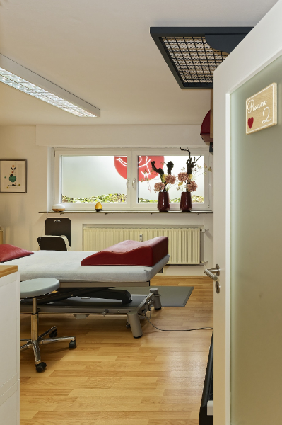 Ein medizinischer Untersuchungsraum mit einer Behandlungsliege für Physiotherapie-Behandlungen, einem Stuhl und Vasen auf der Fensterbank. Die Tür ist offen und ein Schild mit der Aufschrift „Raum 2“ steht darauf.