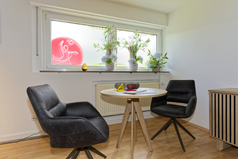 Hier kann man vorab die Physiotherapie-Behandlung besprechen: Zwei schwarze Stühle und ein runder Holztisch mit Notizbüchern, Stiften und Glaswaren stehen in einem hellen Raum, Topfpflanzen auf der Fensterbank.