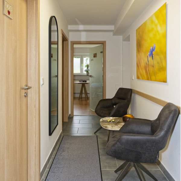 Ein moderner Flur mit zwei grauen Sesseln, einem kleinen runden Tisch, einem großen Spiegel und einer Erinnerungskarte für Ihren Physiotherapie-Termin neben dem leuchtend gelben Blumenfoto an der Wand.