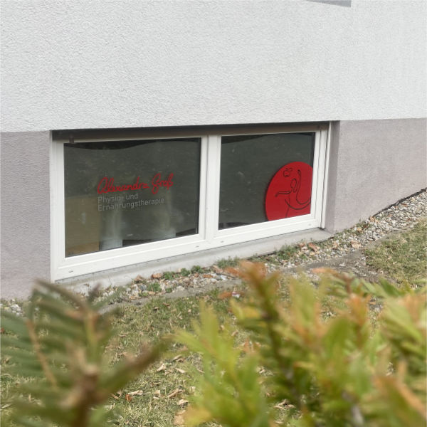Ein Fenster im Souterrain ist mit dem roten Schriftzug Alexandra Groß und dem roten Bildlogo beklebt und so weist so den zu Ihrem nächsten Physiotherapie-Termin.